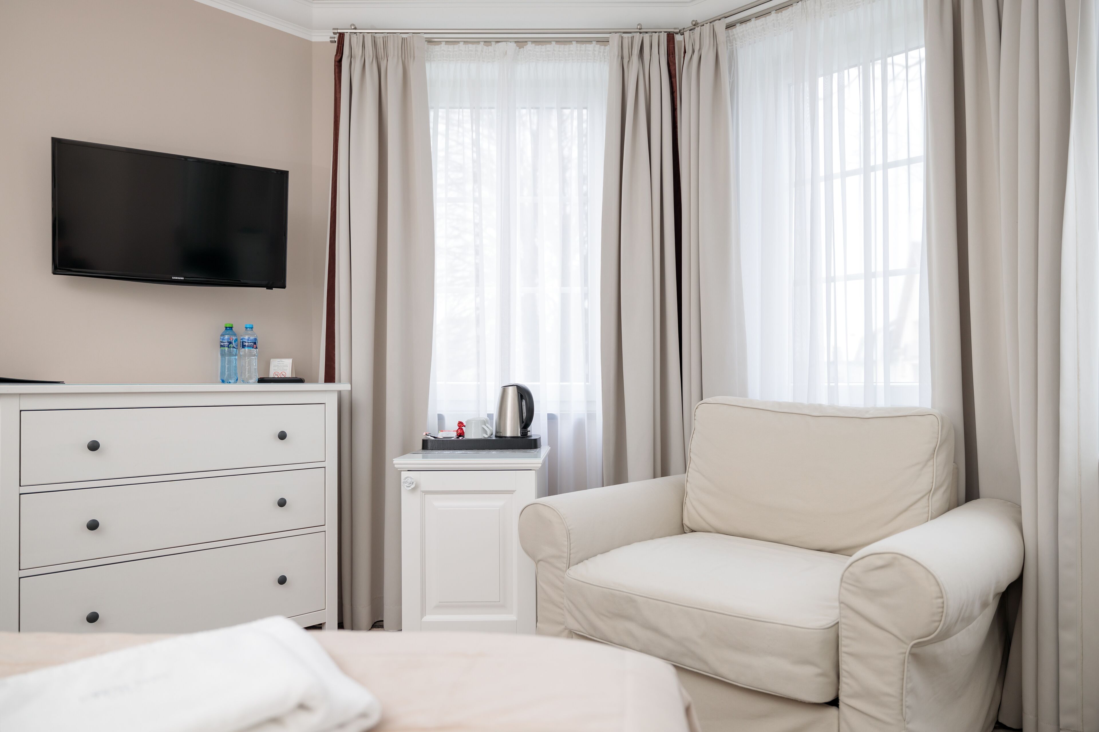 Habitación de hotel con una cama, un tocador blanco, televisor, ventana con cortinas y un sillón color beige.