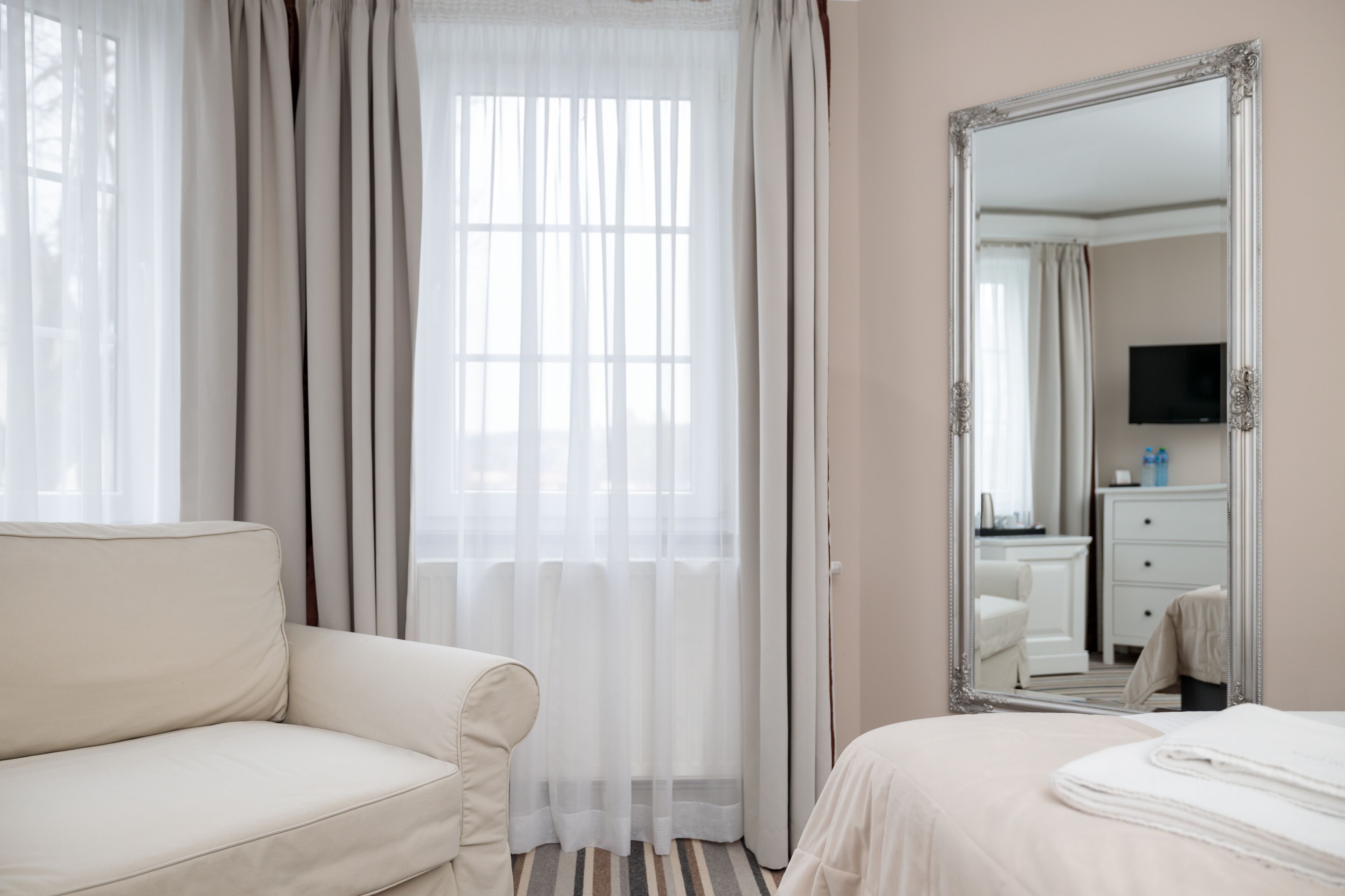 A hotel room with a large mirror, a bed, a sofa, and a window with sheer curtains.