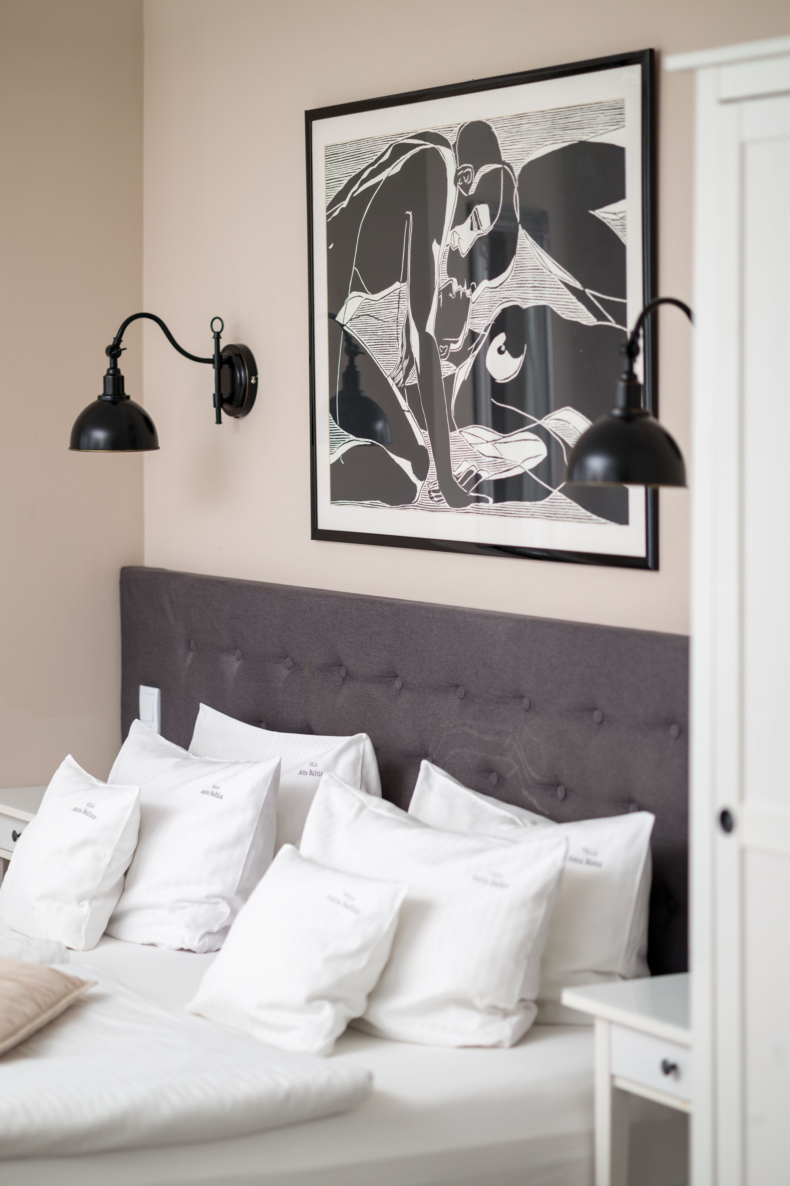 Un lit bien fait, avec une literie blanche et de nombreux oreillers, une tête de lit rembourrée de couleur grise et une œuvre d’art abstraite noire accrochée au mur.