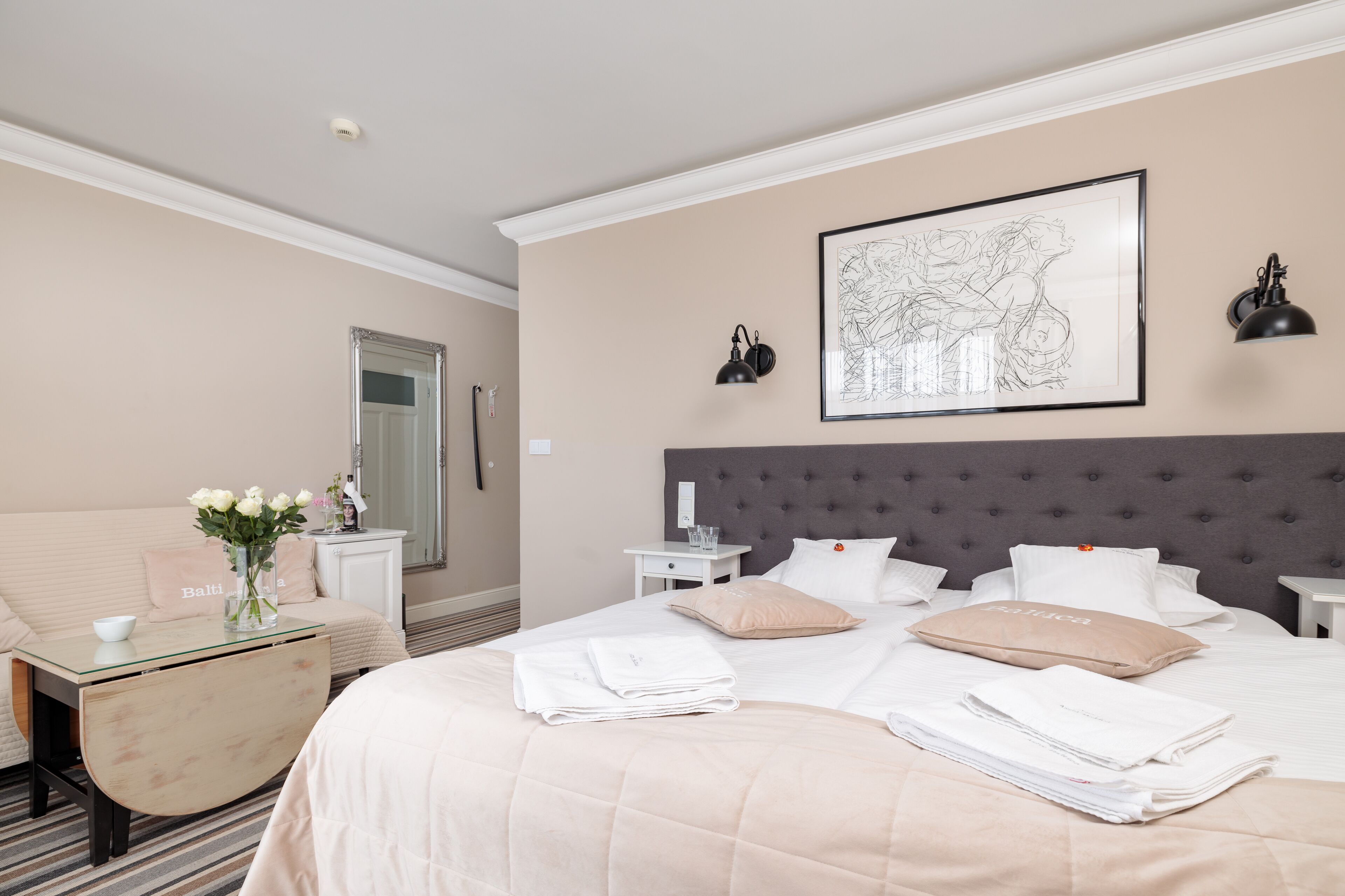 A neatly arranged hotel room with a large bed, a bedside table, a small desk with a vase of flowers, and a framed artwork on the wall.