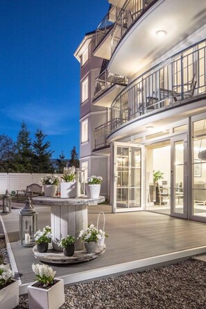 Un immeuble à plusieurs étages doté d’un balcon, d’un espace extérieur aménagé pour s’asseoir et d’un jardin circulaire orné de fleurs.