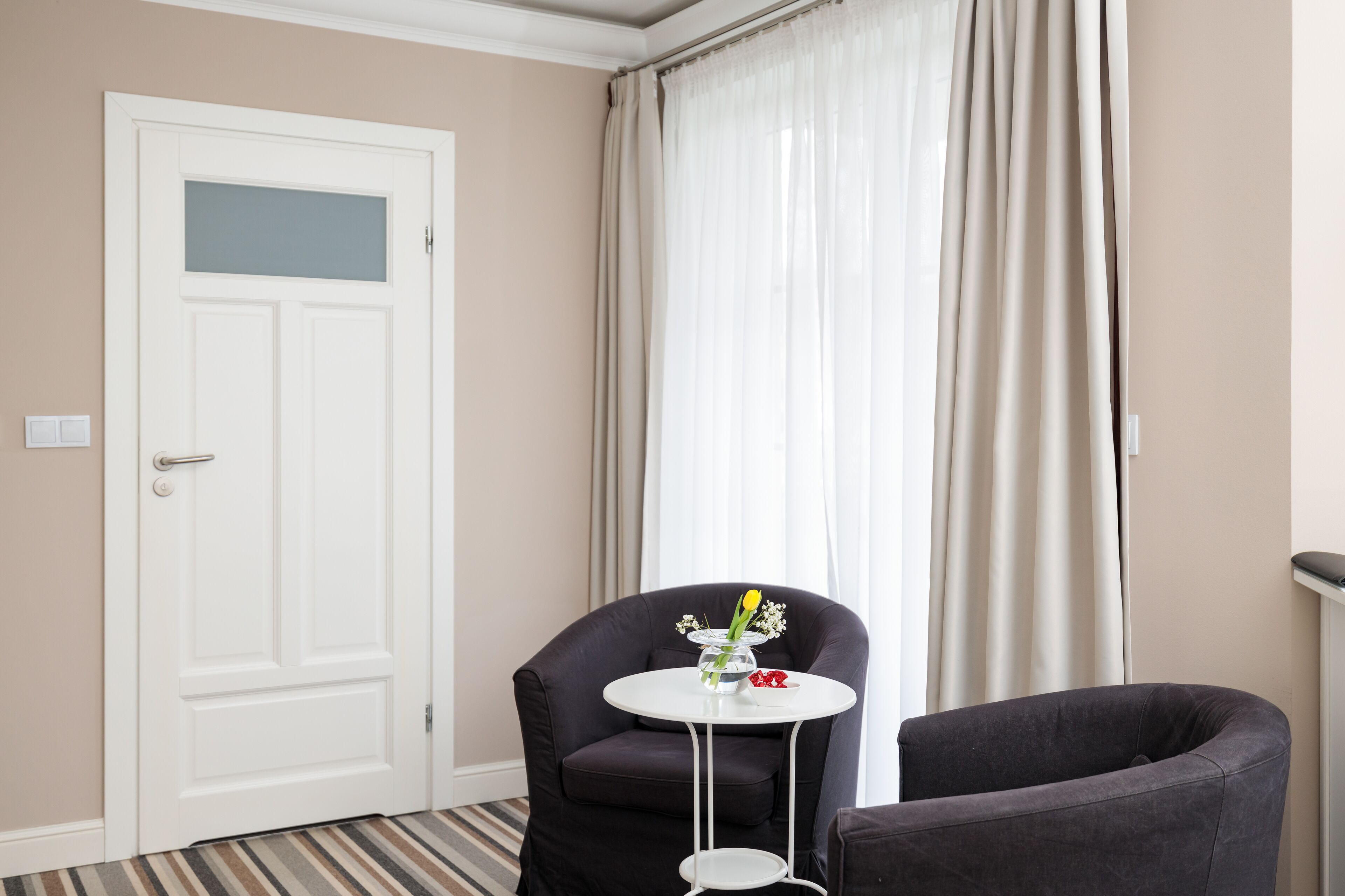 Ett hotellrum med en vit dörr, en svart fåtölj, ett litet runt bord med en vas med blommor och ett stort fönster med tunna, genomskinliga gardiner.