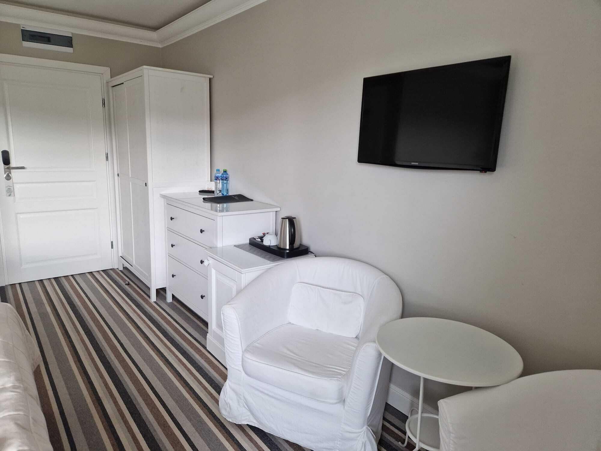 Habitación de hotel con un sillón blanco, una mesita pequeña, un televisor de pantalla plana en la pared, un minibar con tetera y una alfombra a rayas.