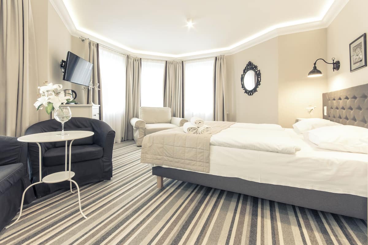 Una habitación de hotel con cama, sofá, una mesita, televisor y ventana con cortinas.