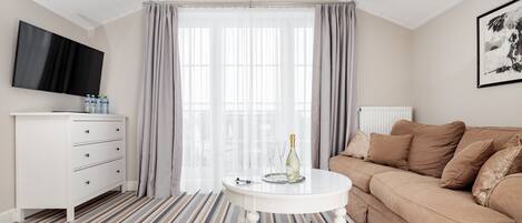 A modern living room with a white sofa, a round coffee table, a flat-screen TV, and a large window with curtains.