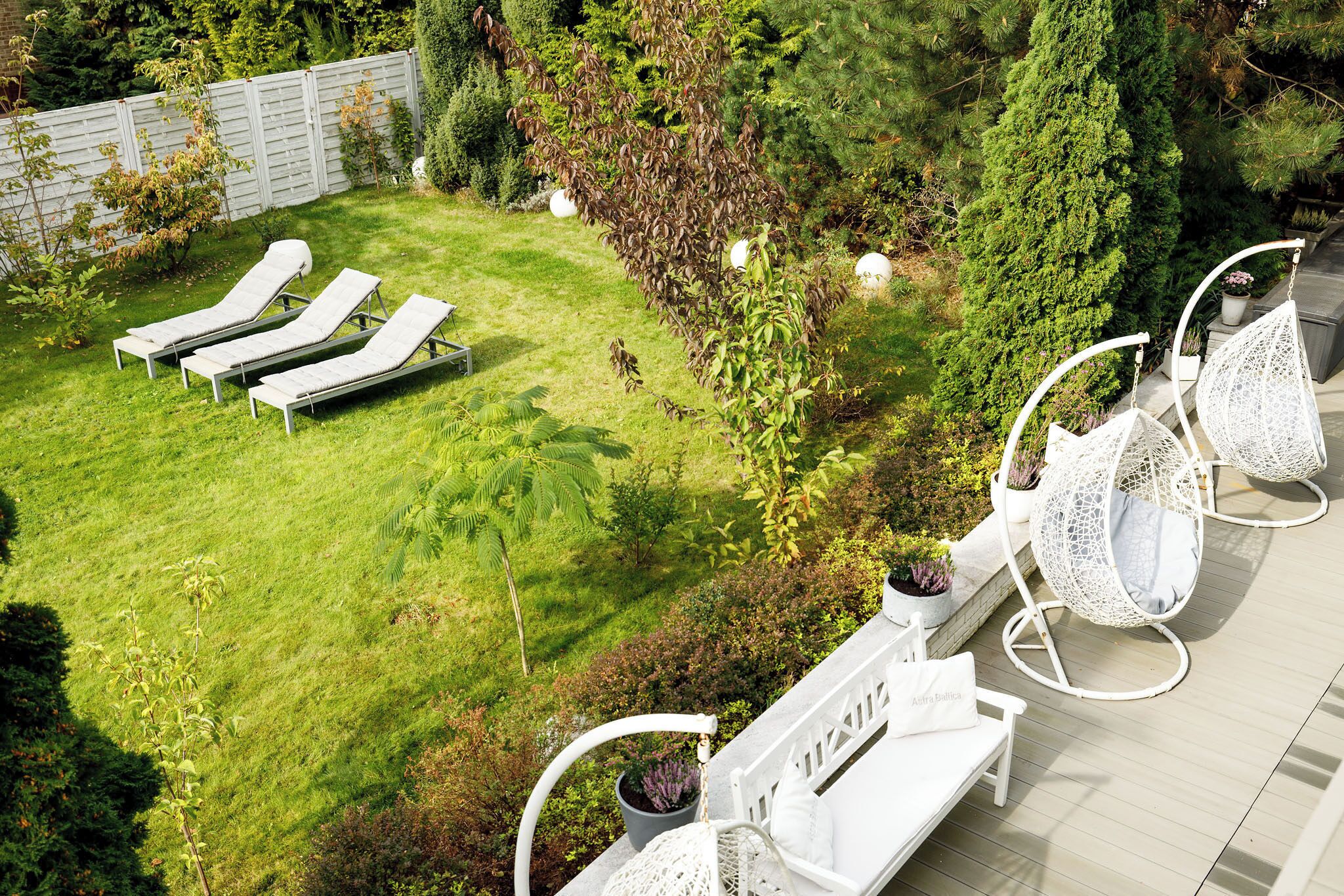 A rooftop garden with lounge chairs, a white fence, and various plants.