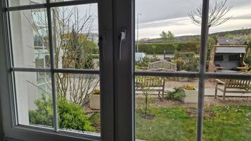 A window with a view of a garden area including a bench, a small structure, and some trees.
