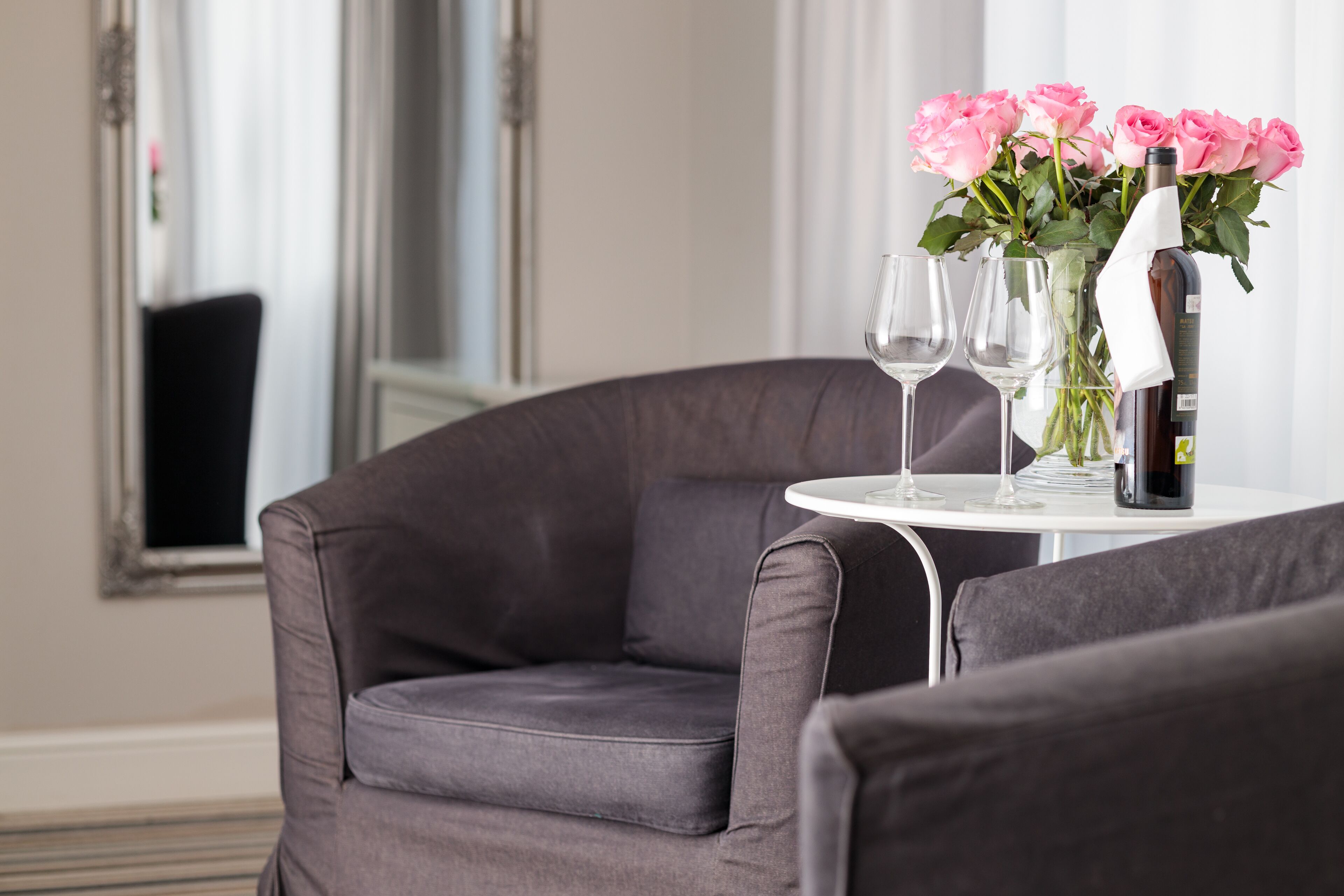 A modern room with a grey sofa, a small round table with a bottle and glasses, and a vase of pink flowers.