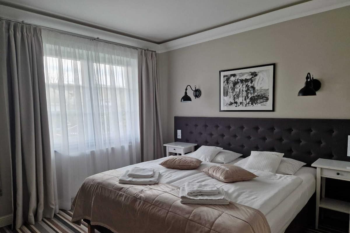 Una habitación de hotel con una cama grande, una mesita de noche, un escritorio, una ventana con cortinas y una lámpara fijada a la pared.