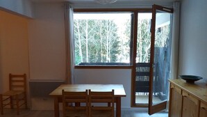 A room with a wooden table and chairs, a window with a view of trees, and a sliding door leading to a balcony.