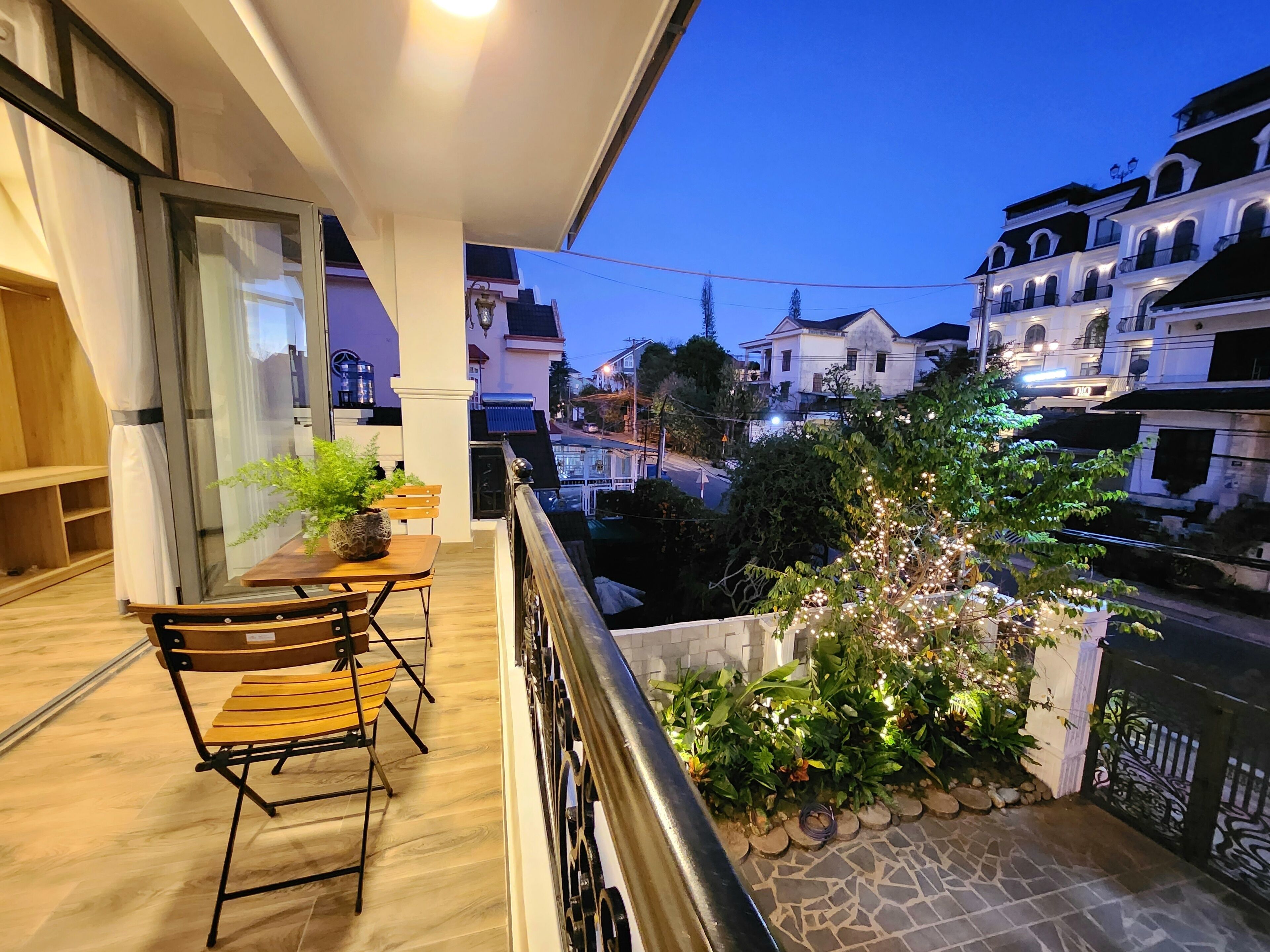 A balcony with a wooden table and chairs, a potted plant, and a view of the cityscape at dusk.
