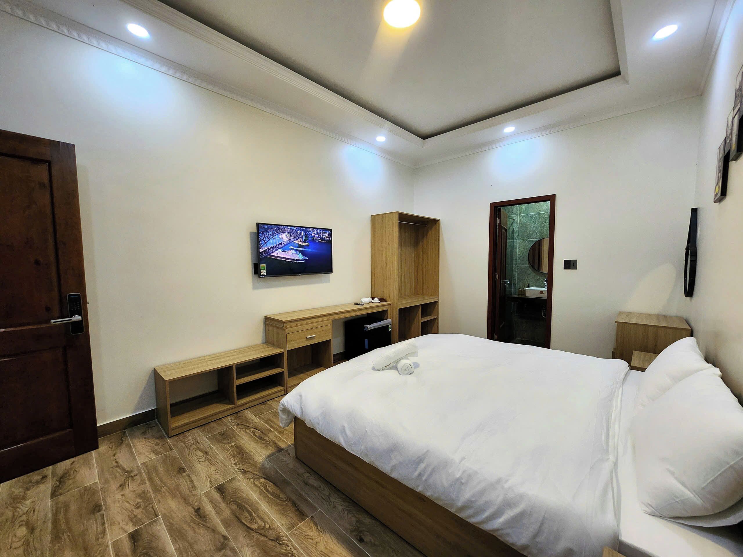 A bedroom with a bed, a TV, a wooden desk, and a bathroom visible through an open door.