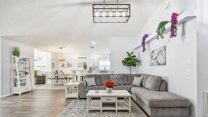 Una sala moderna con un sofá seccional gris, una mesa de centro blanca y un tapete estampado. A la izquierda, hay una estantería blanca con plantas decorativas.