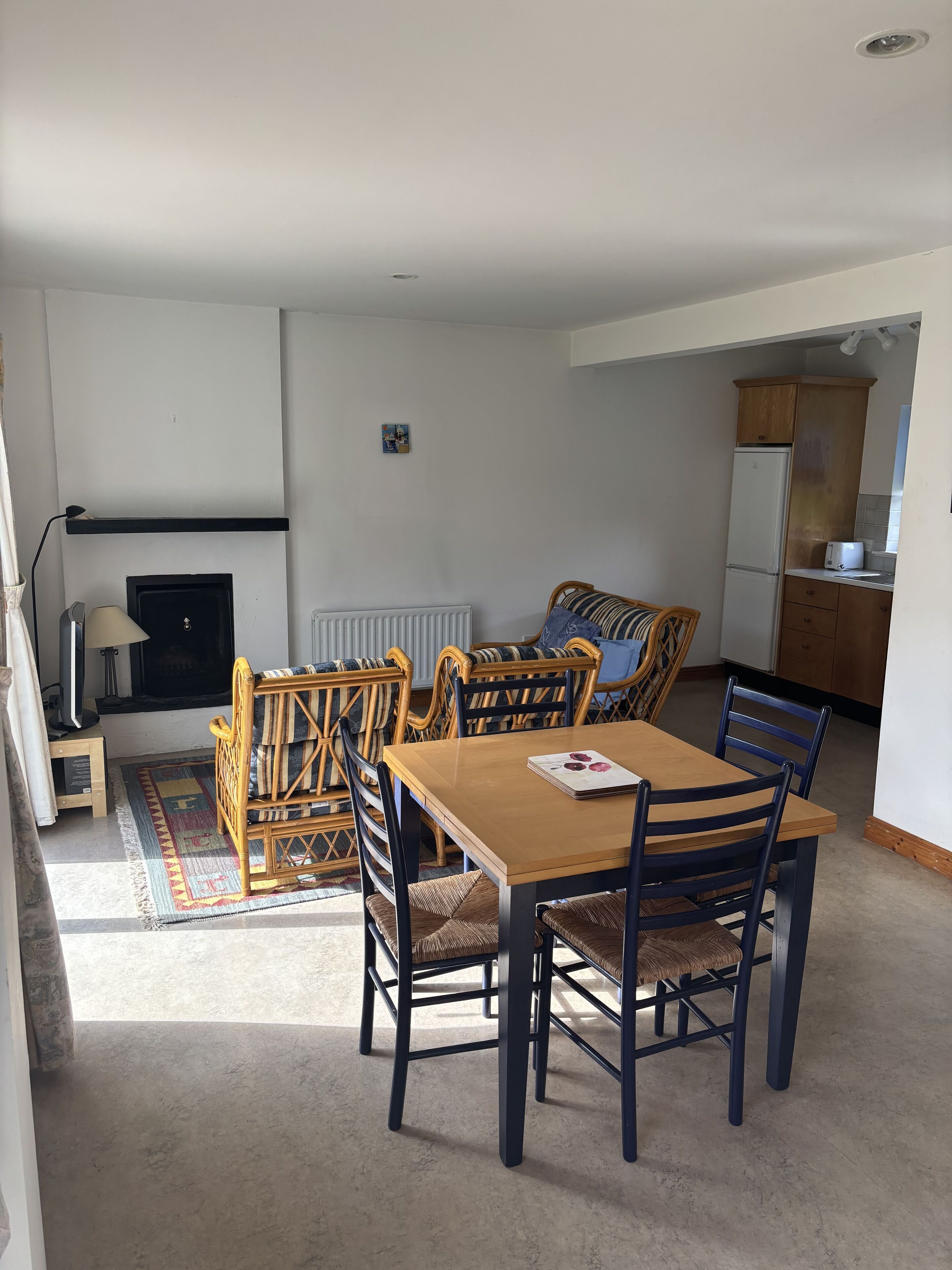 A living room with a wooden dining table, wicker chairs, a fireplace, and a kitchen area with wooden cabinets and a refrigerator.