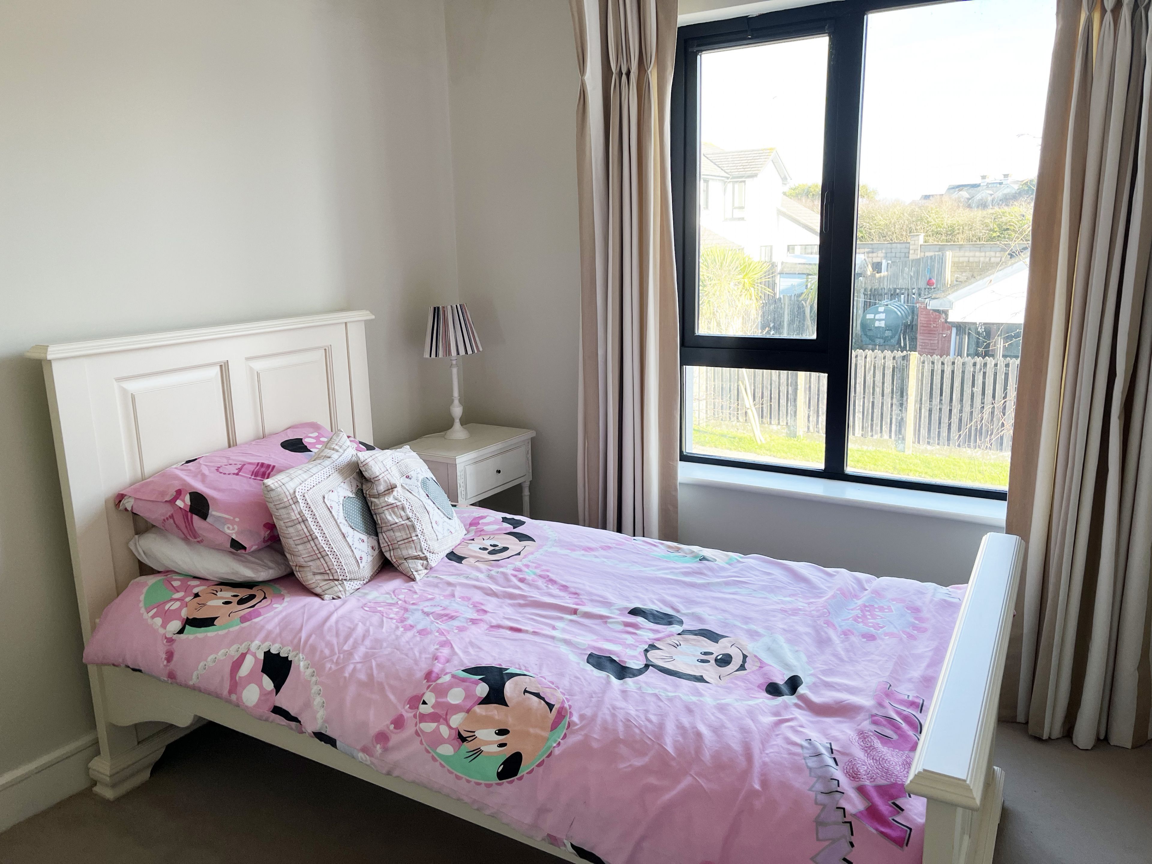 A bedroom with a bed featuring a pink Minnie Mouse themed bedspread, a white headboard, and a nightstand with a lamp.