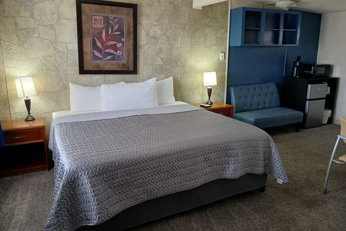 Quarto de hotel com uma cama, duas mesas de cabeceira com abajures, uma poltrona azul, uma pequena escrivaninha com um computador e um espaço com cozinha compacta.