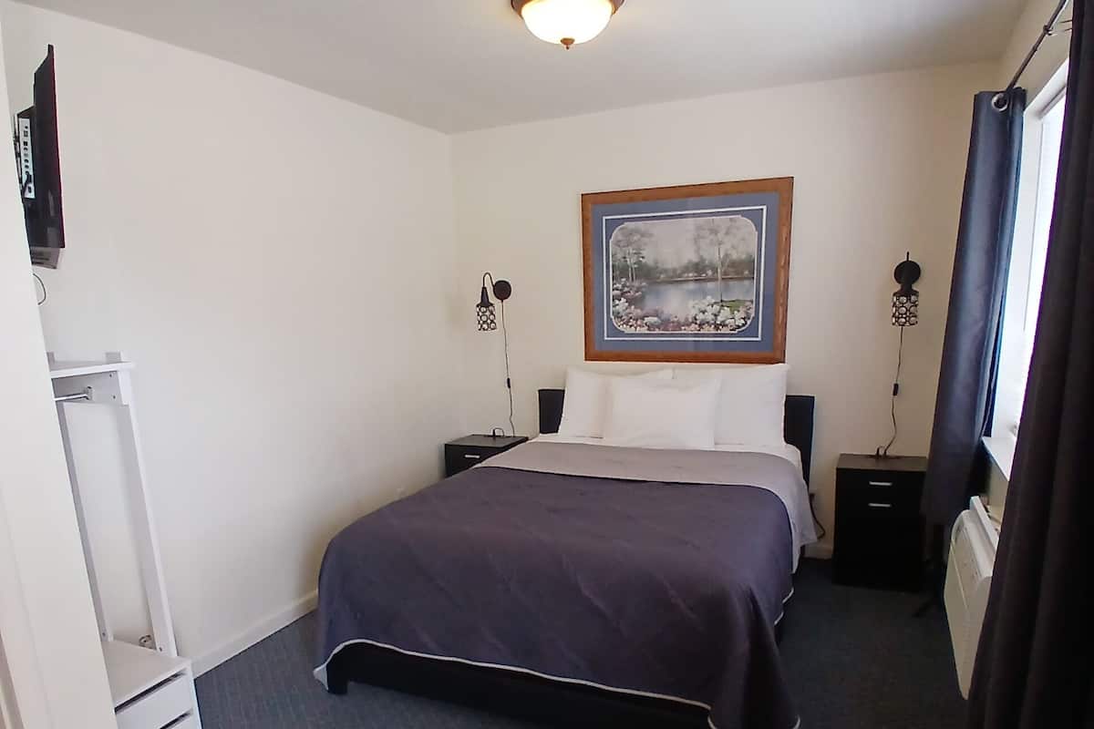 Quarto de hotel com cama, mesas de cabeceira, escrivaninha, cadeira, quadro na parede e luminária no teto.