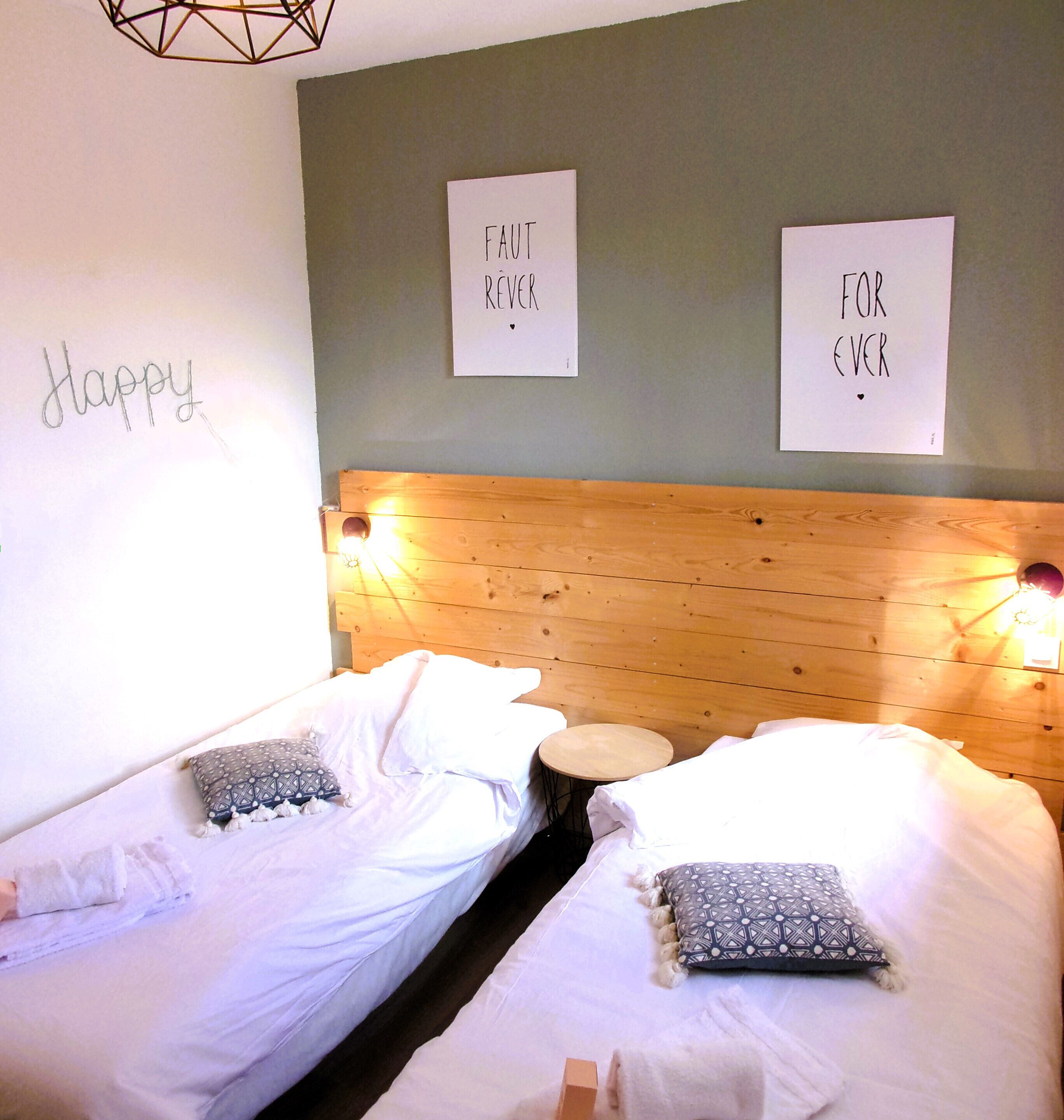 Une chambre avec deux lits, chacun agrémenté de draps blancs et d’oreillers à motifs. Deux panneaux avec du texte sont fixés au mur, et un luminaire aux formes géométriques est suspendu au-dessus des lits.