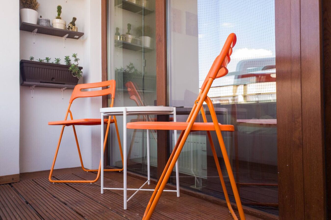 Ein Balkon mit zwei orangefarbenen Stühlen, einem kleinen Tisch, Holzboden und Regalen mit Topfpflanzen.