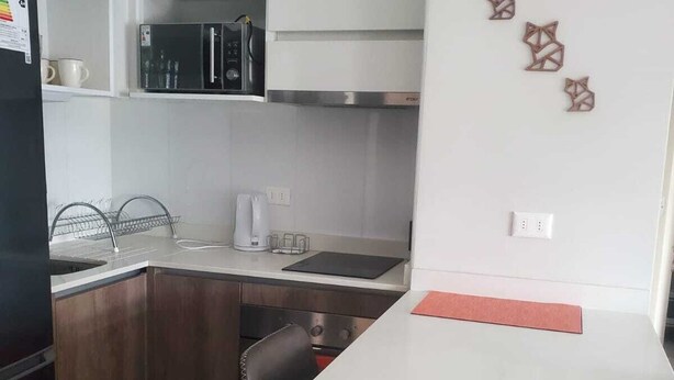 Uma cozinha compacta com armários brancos, uma geladeira preta, micro-ondas e cafeteira. Há peças decorativas na parede e um luminário sobre o balcão.