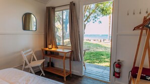 A bedroom with a bed, a desk with a lamp, a chair, a window with curtains, and a view of the beach. - White s Landing Catalina Camp (Avalon)