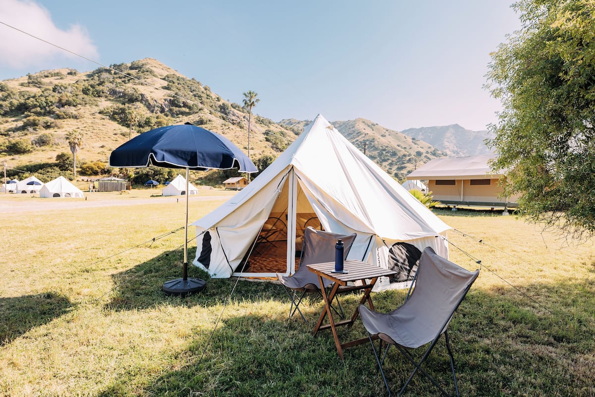 Une tente avec une table et des chaises installées dans un espace herbeux, avec une montagne en arrière-plan.