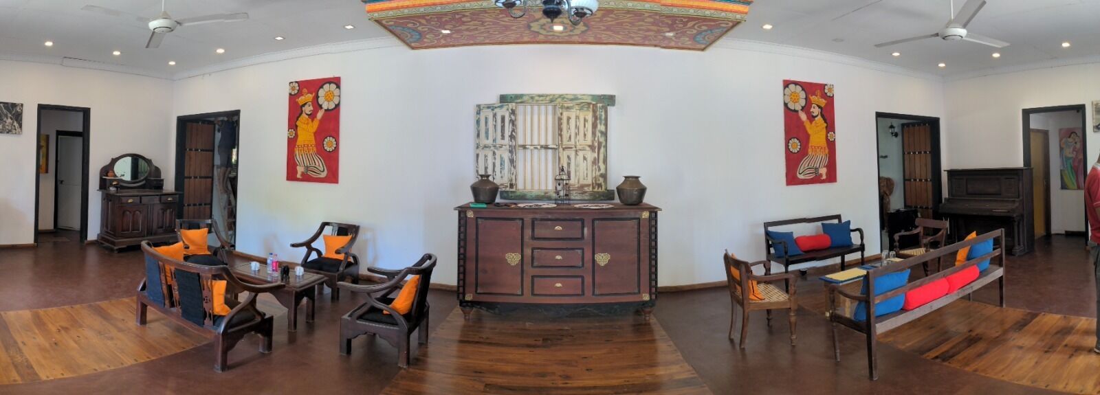 A spacious living room with a wooden floor, a central cabinet, two sofas with colorful cushions, and a ceiling fan.