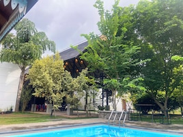 Piscina com borda de pedra, rodeada por árvores e um edifício com telhado escuro e luminárias pendentes.