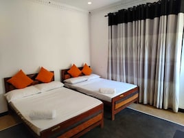 Quarto de hotel com duas camas de solteiro, cada uma com roupa de cama branca e almofadas laranja. O quarto possui cabeceira de madeira, piso escuro e janela com cortinas leves.