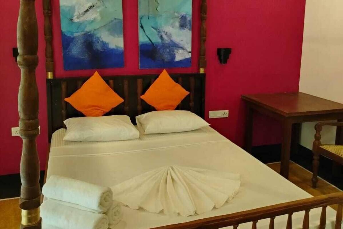 A four-poster bed with white linens and orange pillows, set against a red wall with two abstract paintings.