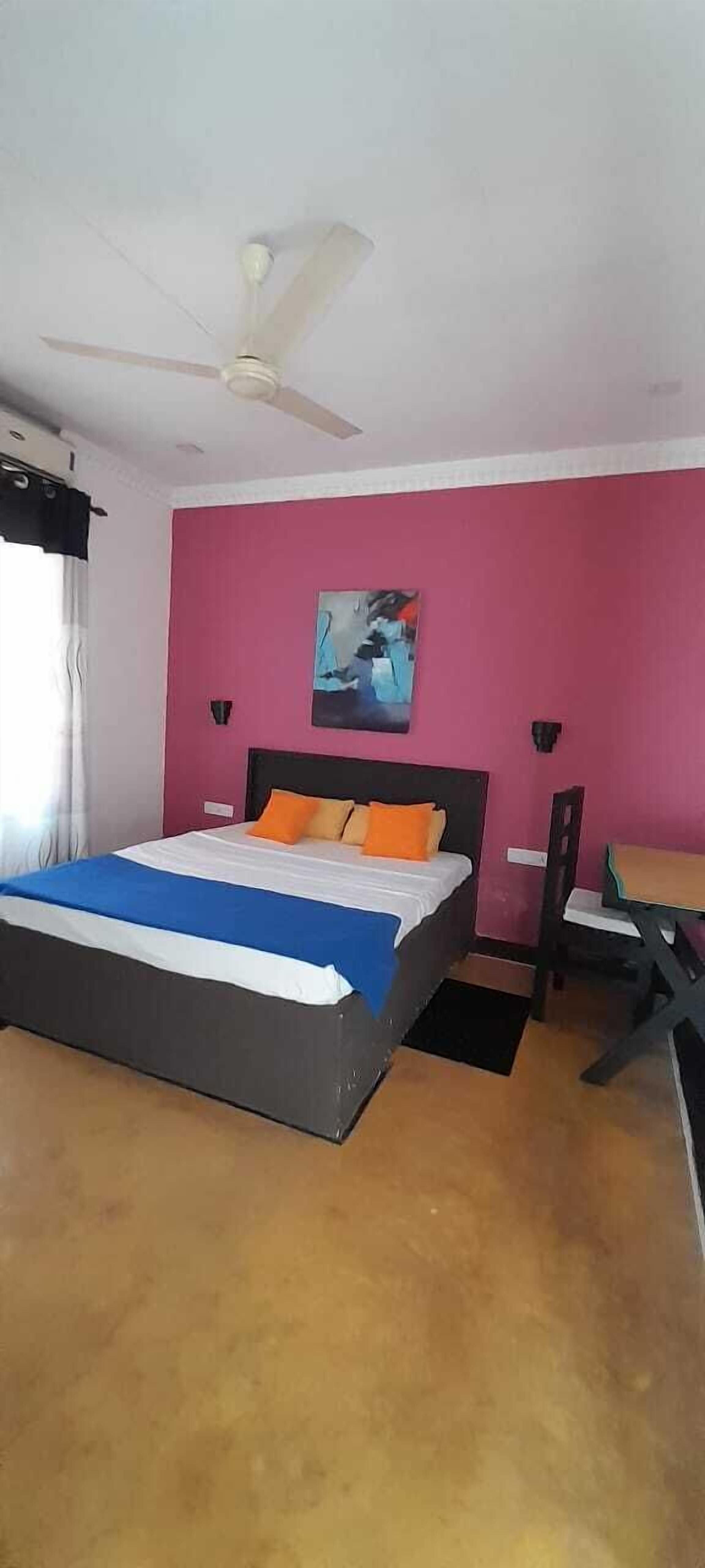 Un dormitorio con cama, escritorio, silla, ventilador de techo y un cuadro en la pared.