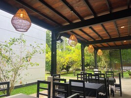 Um pátio externo com mesas e cadeiras, vigas de madeira, luminárias de vime pendentes e plantas.