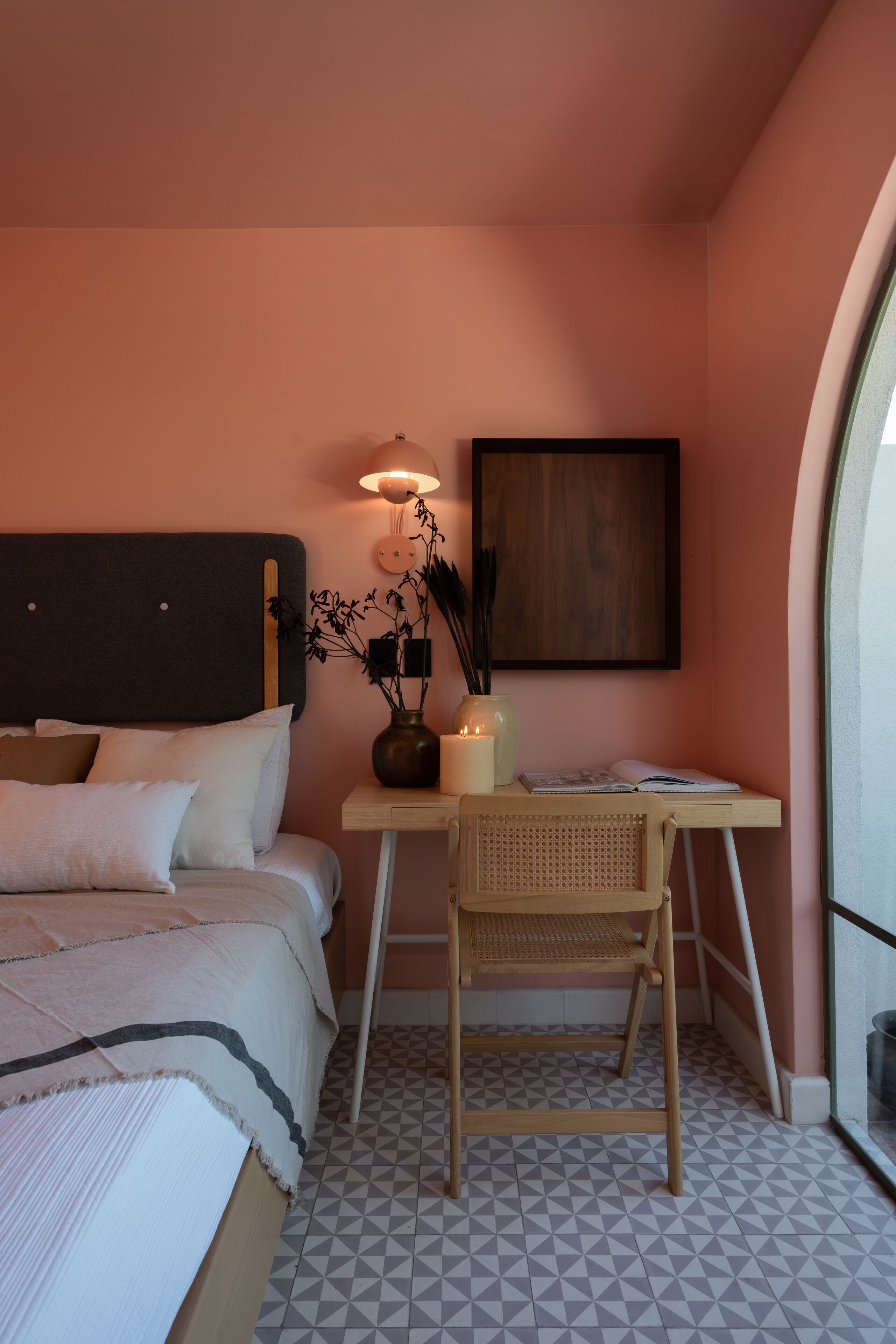 A bedroom with a bed, bedside table, lamp, vase with plants, and a framed picture on the wall.