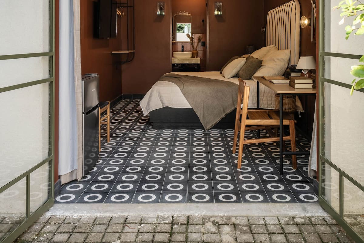 Une chambre à coucher compacte et bien éclairée, avec un lit, une table de chevet et un miroir. La pièce est dotée d’un sol à motifs et d’une petite fenêtre.