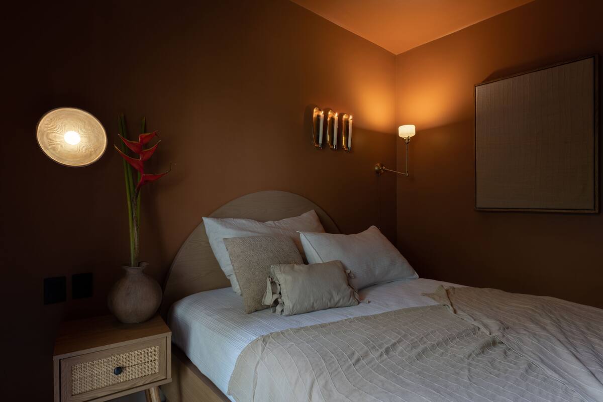 Un lit bien fait, avec une couette blanche et des oreillers, une lampe de chevet, un vase avec des fleurs rouges et un luminaire fixé au mur.