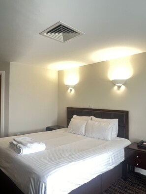 Une chambre d’hôtel avec un grand lit, deux lampes fixées au mur, une grille d’aération au plafond et une table de chevet avec un téléphone.
