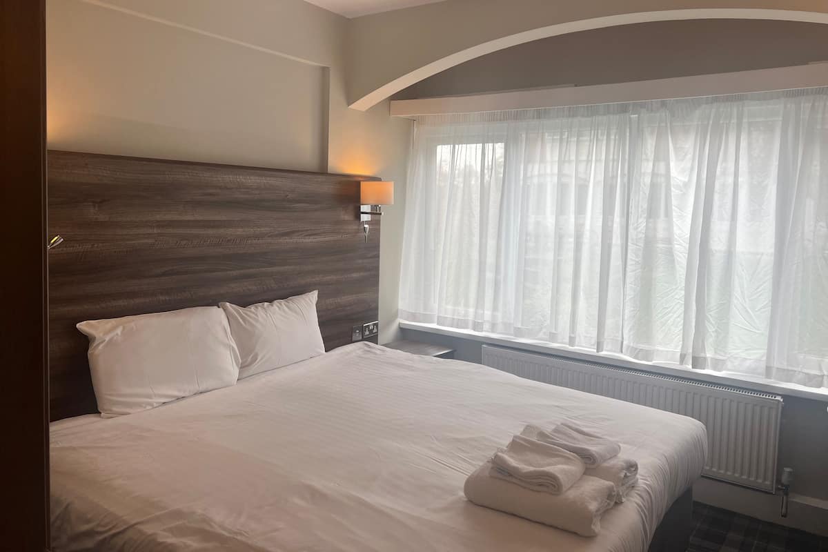 A neatly made bed with white linens, a wooden headboard, a bedside lamp, and a window with sheer curtains.
