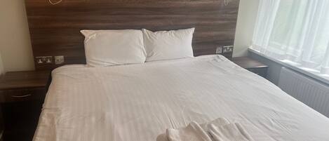 A neatly made bed with white linens, two bedside lamps, and a window with sheer curtains.