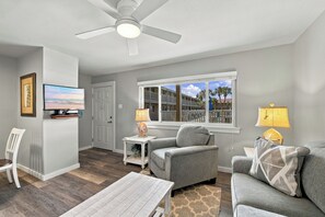 A living room with a ceiling fan, a television, a sofa, a coffee table, and a window with a view of a building. - Roomy 1 BDRM block from the BEACH Large Pool w/ a Private Courtyard (Port Aransas)