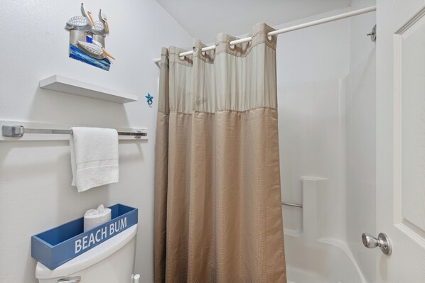 A bathroom with a shower curtain, a towel rack, a toilet paper holder, and a shelf with a towel and a box labeled "BEACH BUM".
