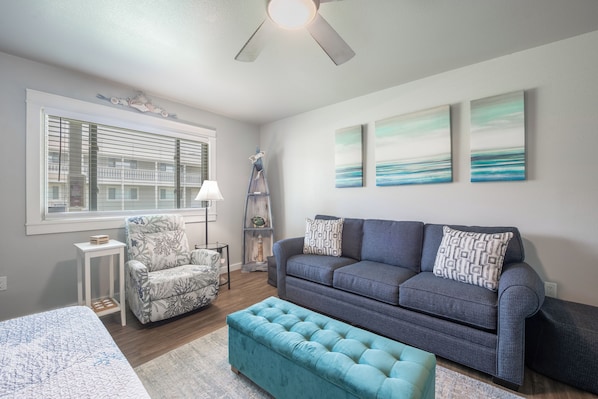 A modern living room with a grey sofa, a patterned armchair, a teal ottoman, and artwork on the walls. - Cozy Studio Large Pool w/ a Private Courtyard and a 5 Min Walk to the Beach (Port Aransas)