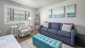 A modern living room with a grey sofa, a patterned armchair, a teal ottoman, and artwork on the walls. - Cozy Studio Large Pool w/ a Private Courtyard and a 5 Min Walk to the Beach (Port Aransas)