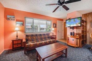 A living room with a sofa, coffee table, and TV. There are two lamps, a ceiling fan, and a bookshelf. - Roomy 1 BDRM Large Pool w/ a Private Courtyard and a 5 Min Walk to the Beach (Port Aransas)