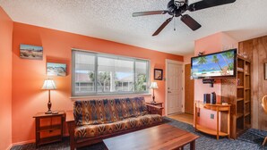 A living room with a sofa, coffee table, and TV. There are two lamps, a ceiling fan, and a bookshelf.