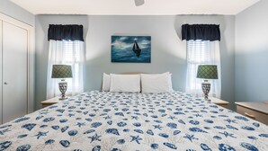 A bedroom with a bed, two nightstands, a window with curtains, a painting of a sailboat, and a patterned bedspread.