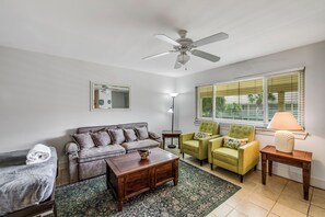 A living room with a ceiling fan, a sofa, two armchairs, a coffee table, a lamp, and a window with blinds. - Roomy 1 BDRM Large Pool w/ a Private Courtyard and a 5 Min Walk to the Beach (Port Aransas)