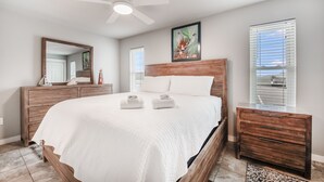 A bedroom with a large bed, wooden headboard, a ceiling fan, a wooden dresser, a mirror, and a window with blinds.