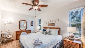 Un dormitorio con cama, ventilador de techo, cómoda, silla, mesita de noche, lámpara, espejo y ventana con persianas.