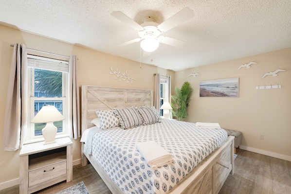 A bedroom with a bed, nightstand, lamp, window, ceiling fan, and wall art.