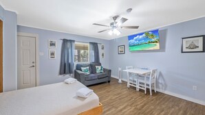 Une chambre à coucher avec un lit, un canapé gris, une table à manger avec des chaises, un ventilateur de plafond et un tableau sur le thème de la plage accroché au mur.